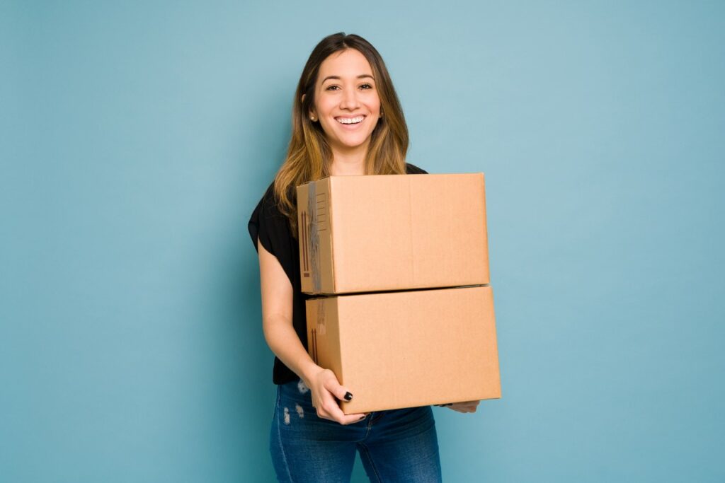 A smiling woman carrying two medium-sized boxes in front of a blue background.