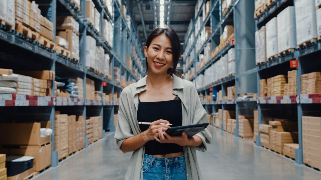 A young entrepreneur holding a tablet smiles for the camera with a warehouse behind. 