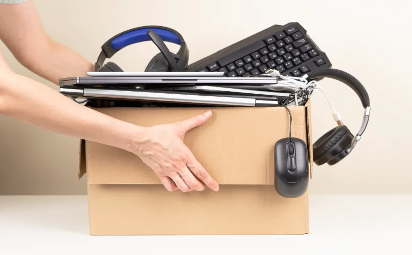 Person carrying a cardboard box filled with electronics, headphones, a keyboard, and cables.